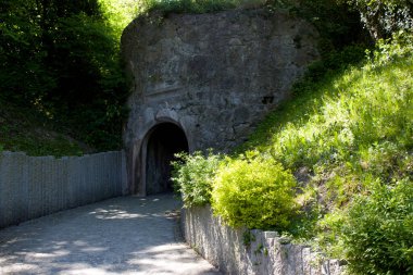 Çimenli bir tepede taş bir duvarla çevrili taş bir tünel girişi. Yeşil alan, doğal ortamda barışçıl ve bir nebze de olsa gizli bir geçit oluşturarak çevresini sarar.