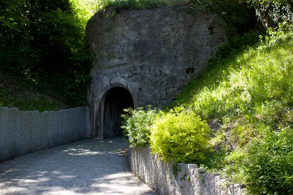 Çimenli bir tepede taş bir duvarla çevrili taş bir tünel girişi. Yeşil alan, doğal ortamda barışçıl ve bir nebze de olsa gizli bir geçit oluşturarak çevresini sarar.