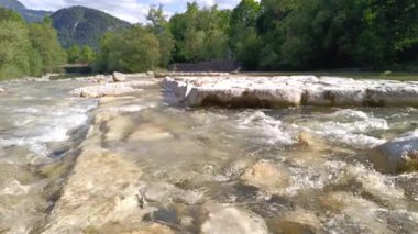 Berrak bir nehirdeki kayaların üzerinden akan temiz su, bereketli ağaçlar ve dağlarla çerçevelenmiş, huzurlu bir açık hava deneyimi yaratıyor..