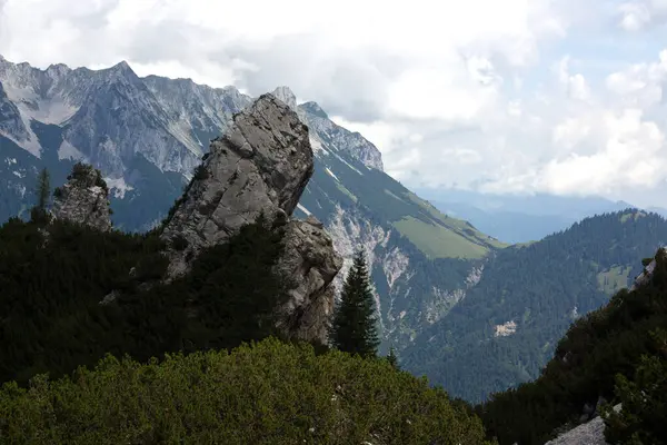 Çarpıcı bir kaya oluşumu, Tirol 'ün yemyeşil arazisinin ortasında belirgin bir şekilde yükselir. Arkaplanda Avusturya 'nın görkemli dağları uzanıyor, zirveleri kısmen bulutlarla örtülü..