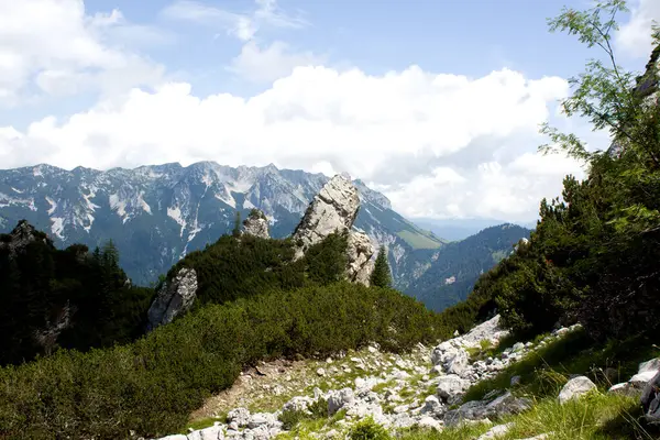 Yuvarlanan tepeler ve yoğun yeşillikler arasında yüksek bir kaya oluşumunun durduğu Avusturya 'nın şaşırtıcı kayalık arazisini deneyin. Arkaplanda parçalı bulutlu gökyüzünün altında görkemli dağlar var.