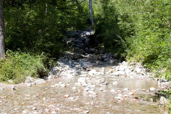 Sakin bir dere yemyeşil bir arazide akar. Sığ sularda ustalıkla istiflenmiş pürüzsüz taşlar parlak güneş ışığı altında huzurlu ve uyumlu bir manzara yaratır..