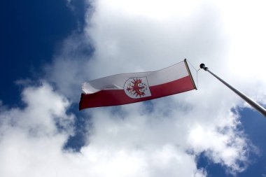 Rüzgarda dalgalanan belirgin bir amblem içeren bir bayrak mavi gökyüzü ve kabarık bulutlara karşı, canlı bir ortamda bölgesel gurur ve kimliği simgeliyor..