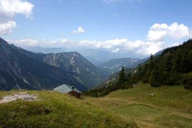 Huzurlu bir dağ manzarası yuvarlanan tepeleri, yemyeşil bir vadiyi ve yumuşak bulutlu parlak mavi gökyüzünün altında yükselen tepeleri gösteriyor. Doğanın içine kurulmuş rahat bir kulübe..