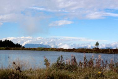 Huzurlu bir göl, yemyeşil çimenler ve kır çiçekleriyle çevriliyken mavi gökyüzünü yansıtır. Uzak dağlar yumuşak, beyaz bulutların altında güzel bir zemin oluşturur..