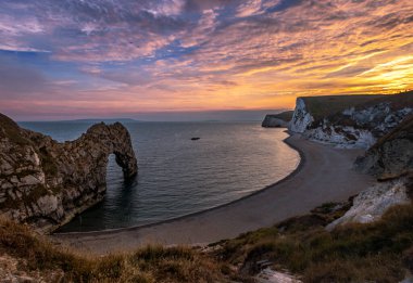 Güneş batıyor ve güzel renkler parçalı bulutlu bir günde Jurassic Sahili 'nde Durdle Door sahilinde yüksek kireçtaşı kayalıkları olan güney İngiltere' de ortaya çıkıyor.. 