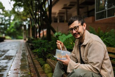 Hippi görünümlü beyaz bir adam öğle yemeğini dışarıda parkta bir beslenme çantasında yiyor. Açık havada yemek, huzur ve yaşam tarzı kavramı için. Yüksek kalite fotoğraf