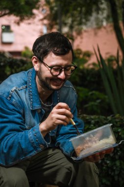 Hippi görünümlü beyaz bir adam öğle yemeğini dışarıda parkta bir beslenme çantasında yiyor. Açık havada yemek, huzur ve yaşam tarzı kavramı için. Yüksek kalite fotoğraf