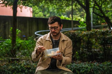 Hippi görünümlü beyaz bir adam öğle yemeğini dışarıda parkta bir beslenme çantasında yiyor. Açık havada yemek, huzur ve yaşam tarzı kavramı için. Yüksek kalite fotoğraf