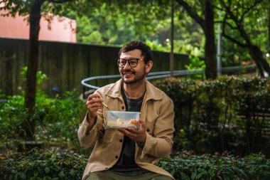 Hippi görünümlü beyaz bir adam öğle yemeğini dışarıda parkta bir beslenme çantasında yiyor. Açık havada yemek, huzur ve yaşam tarzı kavramı için. Yüksek kalite fotoğraf