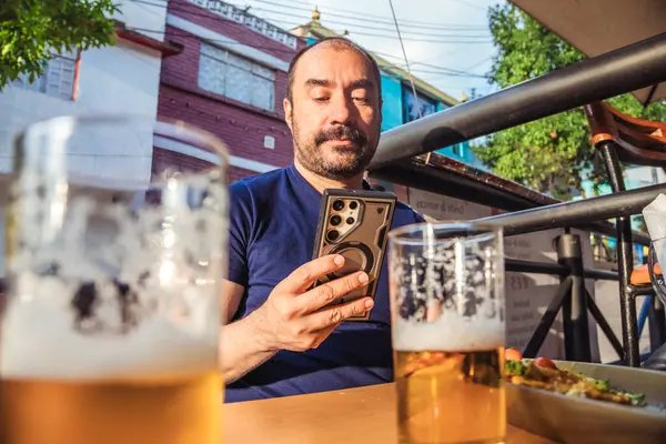 Bir bardak birası olan Latin bir adam kutlama ve yaşam tarzı konsepti için gün batımında bir koloni caddesinde cep telefonuna bakıyor. Yüksek kalite fotoğraf