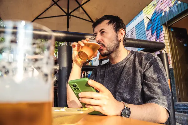 Bir bardak birası olan Latin bir adam kutlama ve yaşam tarzı konsepti için gün batımında bir koloni caddesinde cep telefonuna bakıyor. Yüksek kalite fotoğraf