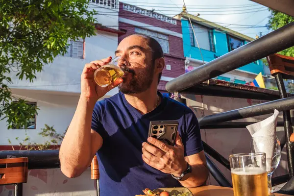 Bir bardak birası olan Latin bir adam kutlama ve yaşam tarzı konsepti için gün batımında bir koloni caddesinde cep telefonuna bakıyor. Yüksek kalite fotoğraf