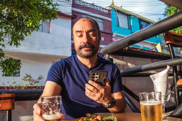 Bir bardak birası olan Latin bir adam kutlama ve yaşam tarzı konsepti için gün batımında bir koloni caddesinde cep telefonuna bakıyor. Yüksek kalite fotoğraf
