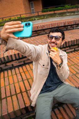 Sakallı ve gözlüklü bir adam dışarıda tuttuğu şeftaliyi yerken selfie çekiyor. Şeftali ayı kavramı, sağlıklı yiyecekler, sağlık ve teknoloji. Yüksek kalite fotoğraf