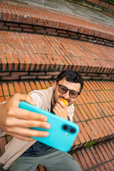Sakallı ve gözlüklü bir adam dışarıda tuttuğu şeftaliyi yerken selfie çekiyor. Şeftali ayı kavramı, sağlıklı yiyecekler, sağlık ve teknoloji. Yüksek kalite fotoğraf
