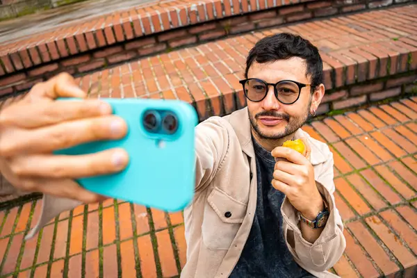 Sakallı ve gözlüklü bir adam dışarıda tuttuğu şeftaliyi yerken selfie çekiyor. Şeftali ayı kavramı, sağlıklı yiyecekler, sağlık ve teknoloji. Yüksek kalite fotoğraf