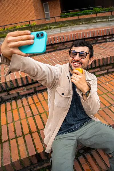 Sakallı ve gözlüklü bir adam dışarıda tuttuğu şeftaliyi yerken selfie çekiyor. Şeftali ayı kavramı, sağlıklı yiyecekler, sağlık ve teknoloji. Yüksek kalite fotoğraf