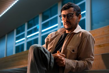 Merdivenlerde tek başına oturan genç bir adam gece hayatı ve moda konsepti için renkli ışıklarla Bogota 'da poz veriyor. Yüksek kalite fotoğraf
