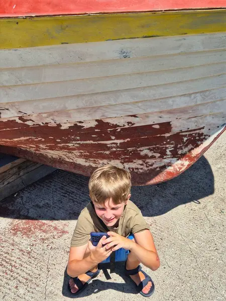 Elinde alet olan bir çocuk eski bir teknenin altında saklanıyor. Sahilde oyun zamanı.