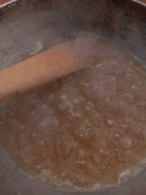 Arka bahçedeki kömür tenceresinde metal tavada domates yahnisi hazırlıyorum. Domates yahnisini pişirirken buharla pişirmenin ilk aşaması, karıştırılmış soğan stoğunu, sıcak yağda pişirilmiş ahşap spatulayla karıştırmak..