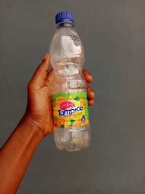 Kumasi, Gana - 26 Şubat 2025: Dişi Afrikalı bir el 500 ml boş şeffaf plastik tampico içecek şişesini kül zemininde tutuyor. İçtikten sonra. Dünya Çevre Farkındalığı Günü.