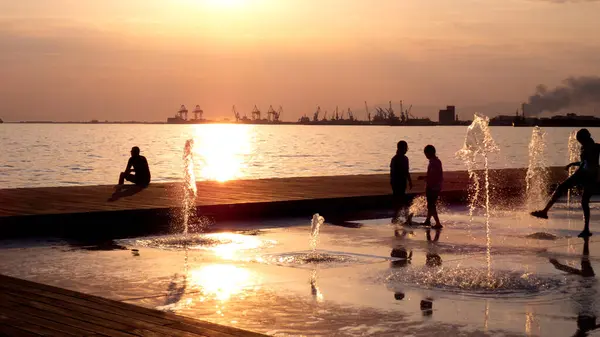 Gün batımında denize bakan şehir çeşmelerinde oynayan çocuklar, altın ışıkla yıkanan sahil şehri gezinti güvertesi, su sıçramaları ve siluetler neşeli bir yaz ortamı yaratır..