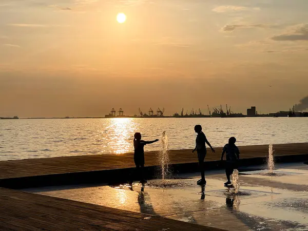 Deniz kenarındaki çeşmede oynayan çocuklar, yaz eğlencesi, su sıçratma, kaarefree anları, deniz kenarındaki mutluluklar, güneşli günler, açık hava aktiviteleri, aile tatili, gülen çocuklar, neşeli sahneler.