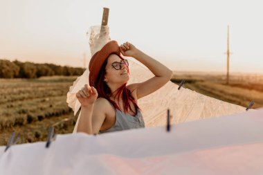 Gözlüklü ve şapkalı gülümseyen kız yazın tarlada beyaz çarşaf asıyor.