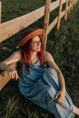 Gözlüklü, şapkalı, gülümseyen kız çitin yanındaki çimlerde oturuyor.