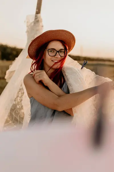 Gözlüklü ve şapkalı gülümseyen kız yazın tarlada beyaz çarşaf asıyor.