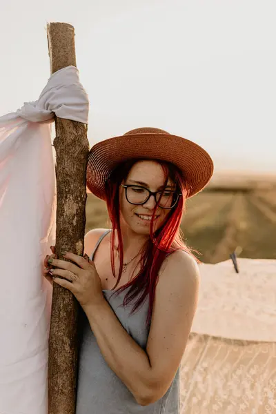 Gözlüklü ve şapkalı gülümseyen kız yazın tarlada beyaz çarşaf asıyor.