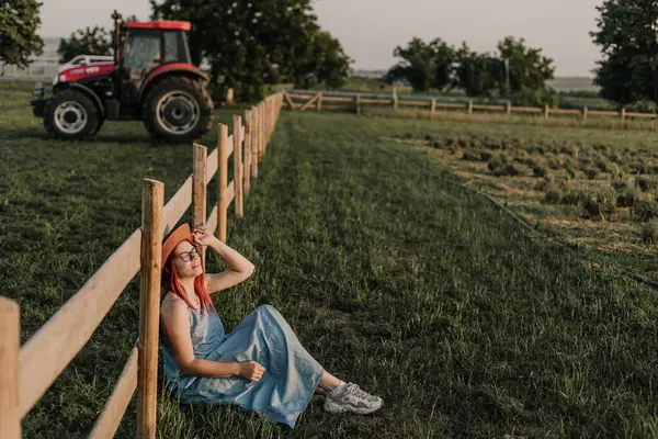 Gözlüklü, şapkalı, gülümseyen kız çitin yanındaki çimlerde oturuyor.