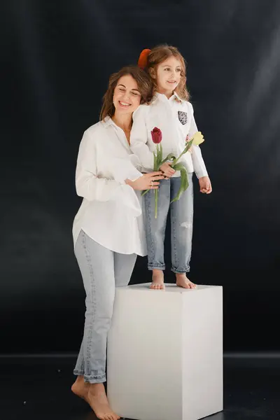 Genç bir kadın ve kızı beyaz tişörtlü ve kotlu koyu arkaplanda bir buket kırmızı çiçekle