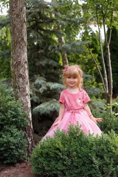 Kıvırcık sarı saçlı, pembe elbiseli küçük kız ağacın yanındaki bankta oturuyor.