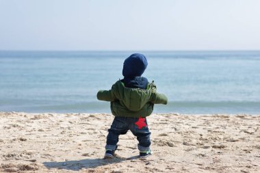 Sırt çantasıyla sahilde duran ve denize bakan çocuk.