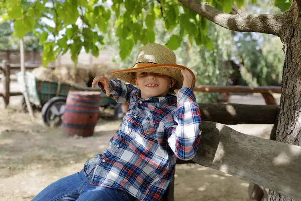 Şapkalı ve kovboylu çocuk ağacın yanında oturuyor.