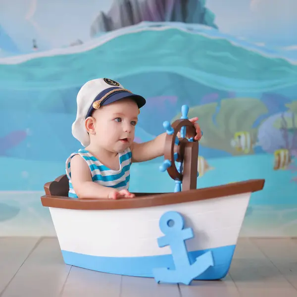 Mavi şapkalı küçük bebek ve denizci gömleği mavi bir denizi ve mavi şapkalı beyaz oyuncağı olan eski bir ahşap teknede oturuyor.