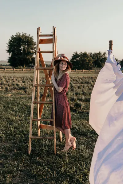 Yazın gün batımında tahta bir merdivenin yanındaki tarlada şapkalı genç bir kız.