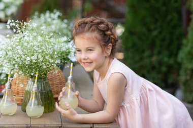 Bahçede kafasında saç örgüsüyle güzel bir kız çiçeklerin arasında yazın limonata içiyor.