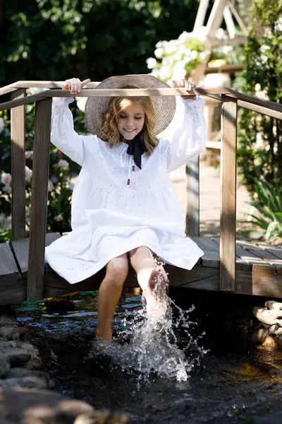 Beyaz elbiseli ve şapkalı güzel kız yazın bahçede tahta köprüde oturuyor.