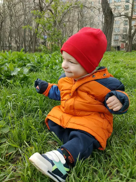 Kırmızı şapkalı ve turuncu ceketli küçük çocuk yeşil çimenlerde oturuyor.