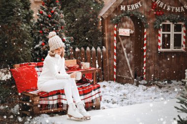 Beyaz kazaklı, beyaz pantolonlu ve kışın patenli bej şapkalı mutlu güzel kız bir fincan kakao ile bankta oturuyor.