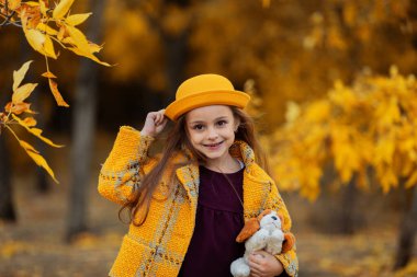 Sonbahar parkında sarı montlu ve sarı şapkalı güzel küçük kız.