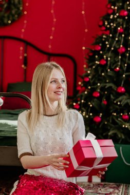 Beyaz elbiseli, sarı saçlı güzel bir kız Noel ağacının yanındaki bir odada hediyelerle oturuyor.