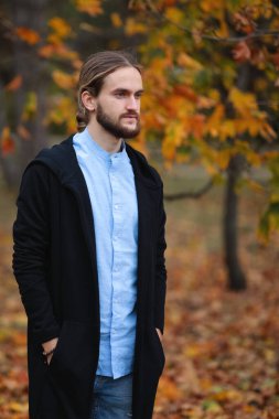 Güz parkında sakallı, uzun saçlı, siyah ceketli ve kot pantolonlu yakışıklı bir genç.