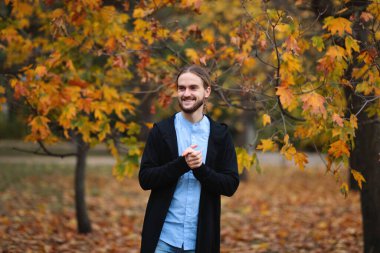 Güz parkında sakallı, uzun saçlı, siyah ceketli ve kot pantolonlu yakışıklı bir genç.