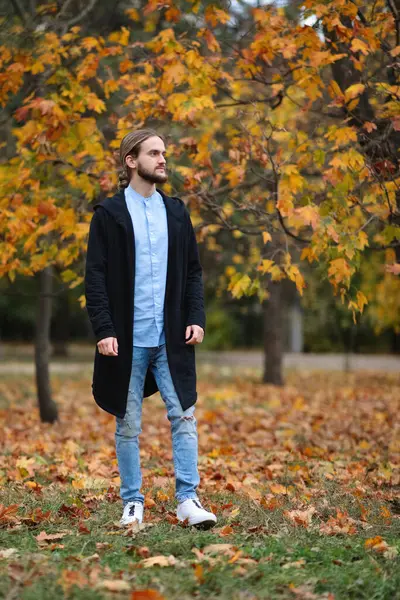 Güz parkında sakallı, uzun saçlı, siyah ceketli ve kot pantolonlu yakışıklı bir genç.