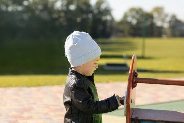 Gri şapkalı ve siyah deri ceketli küçük mutlu çocuk sonbaharda dışarıda oyun alanında oynuyor.