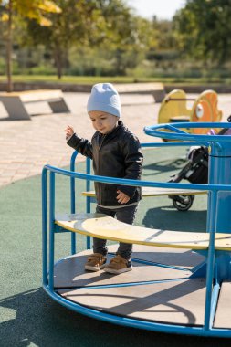 Gri şapkalı ve siyah deri ceketli küçük mutlu çocuk sonbaharda dışarıda oyun alanında oynuyor.